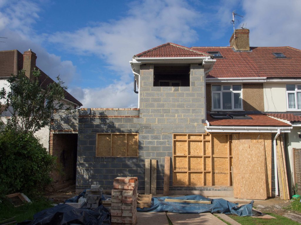 planning permission to replace windows | Black Country Glazing Front view of a house under construction with gray block walls, a partially completed second level, and stacks of bricks on the yard blue tarp